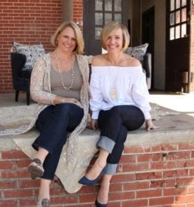 Bekel Designers Beth and Kelly. Two white women with blonde hair sitting in front of a brick wall together.